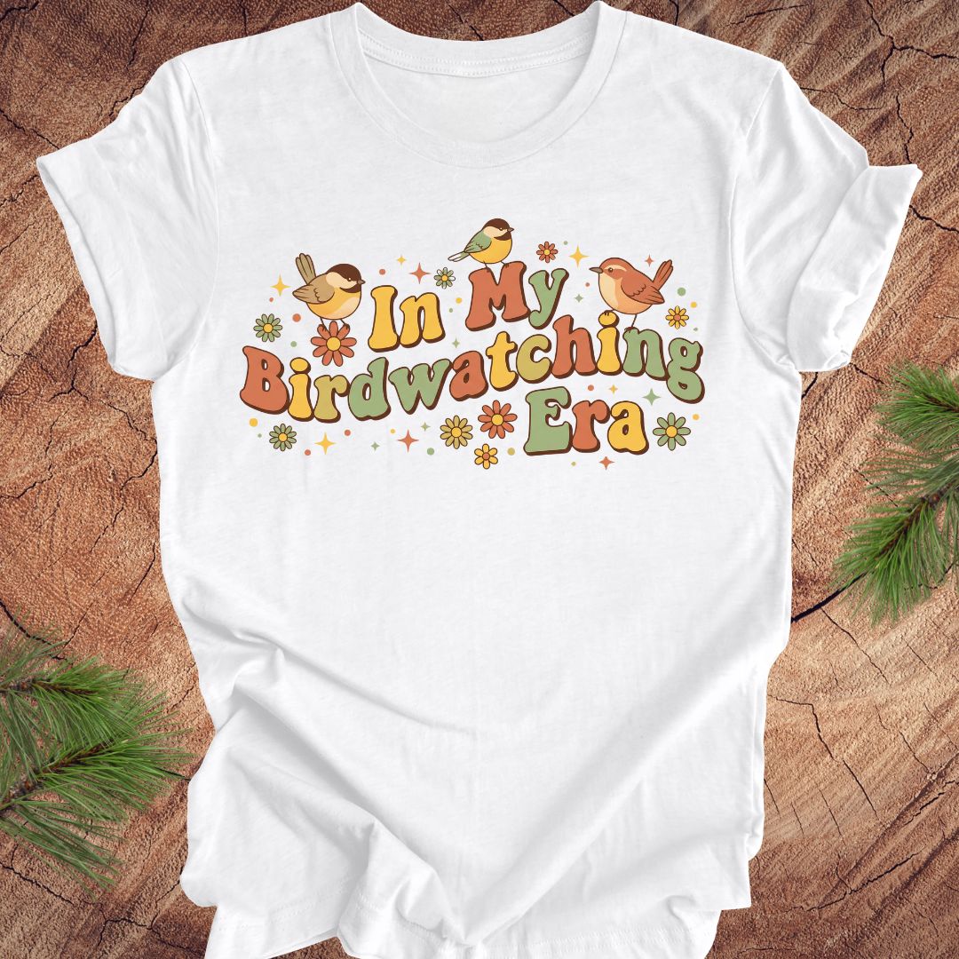 White t-shirt with 'In My Birdwatching Era' text and bird illustrations on a wooden surface.