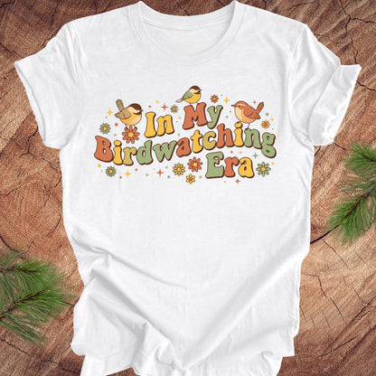 White t-shirt with 'In My Birdwatching Era' text and bird illustrations on a wooden surface.