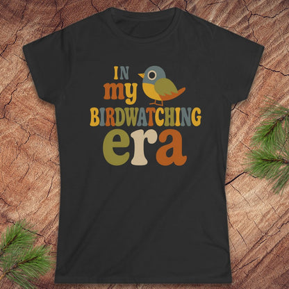 A black short-sleeve graphic t-shirt laid flat on a textured wood background with pine branches in the corners. The graphic reads 'IN my BIRDWATCHING era' in a retro multicolor font, accompanied by a small bird illustration.