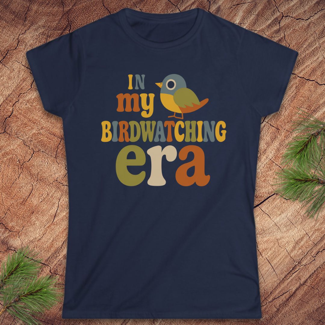 A navy blue short-sleeve graphic t-shirt laid flat on a textured wood background with pine branches in the corners. The shirt features a colorful retro-style graphic that reads 'IN my BIRDWATCHING era' with a small bird illustration.