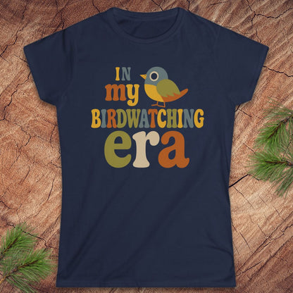 A navy blue short-sleeve graphic t-shirt laid flat on a textured wood background with pine branches in the corners. The shirt features a colorful retro-style graphic that reads 'IN my BIRDWATCHING era' with a small bird illustration.
