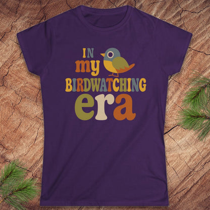 A purple short-sleeve crewneck t-shirt with a retro-style birdwatching graphic reading 'IN my BIRDWATCHING era' in multicolor letters, featuring a small bird illustration. The shirt is laid flat on a textured wood background with pine branches in the corners.