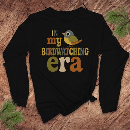 Black long sleeve tshirt with 'In my BIRDWATCHING era' text and bird graphic on a wooden surface.
