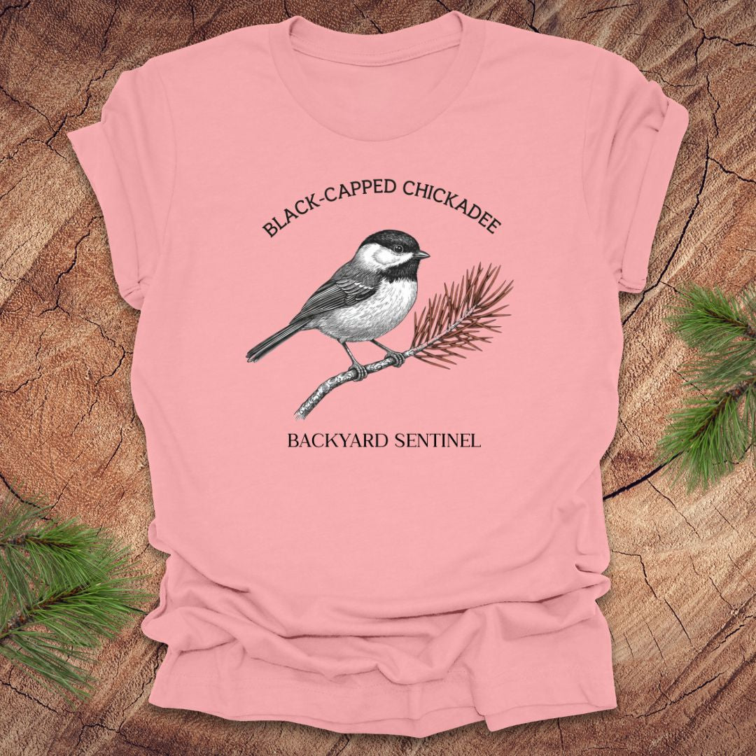 Pink t-shirt with black-capped chickadee design on a wooden surface with pine branches.