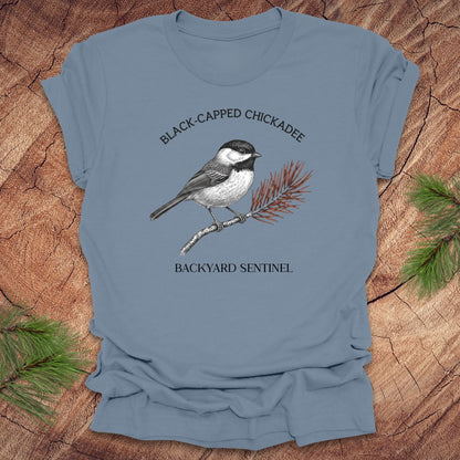 Blue t-shirt with a black-capped chickadee graphic and text on a wooden surface.