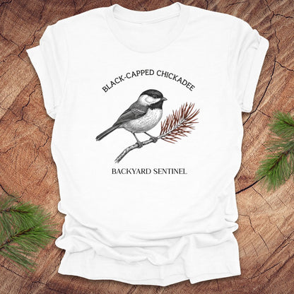 White t-shirt with black-capped chickadee design on a wooden surface