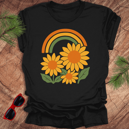 Black t-shirt with a rainbow and sunflower design on a wooden surface with sunglasses and greenery.
