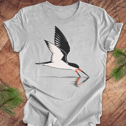 A light gray crewneck t-shirt featuring a graphic of a black and white bird with an orange beak, shown flat on a rustic wood background with pine branches.