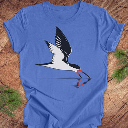 A blue short-sleeve crewneck t-shirt featuring a graphic of a black and white bird with a red-orange beak, laid flat on a rustic wood background with pine branches in the corners.