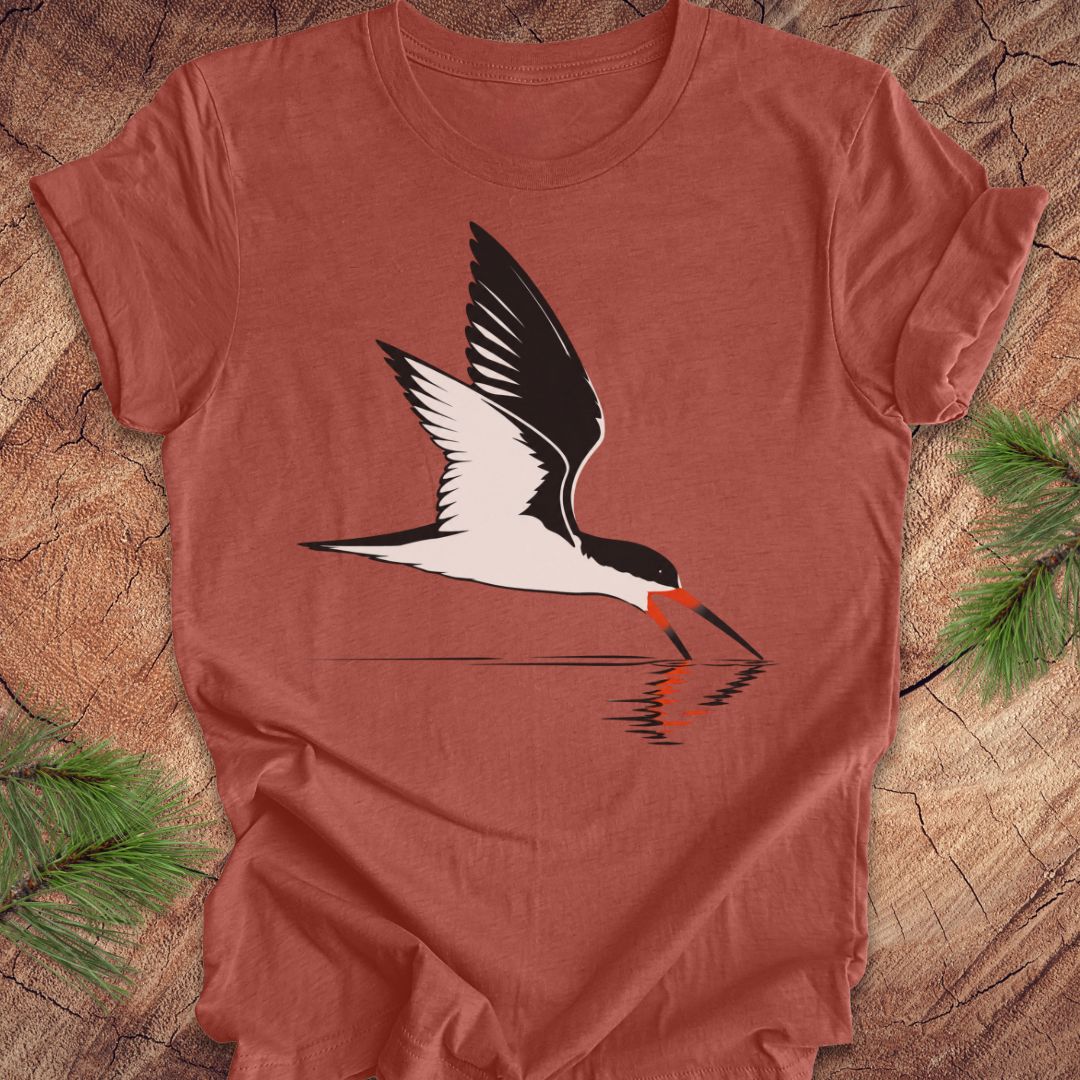 A heathered terracotta short-sleeve t-shirt featuring a graphic of a black and white skimmer bird with a red-orange beak, shown flat on a rustic wood background with pine branches.