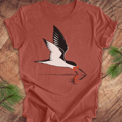 A heathered terracotta short-sleeve t-shirt featuring a graphic of a black and white skimmer bird with a red-orange beak, shown flat on a rustic wood background with pine branches.
