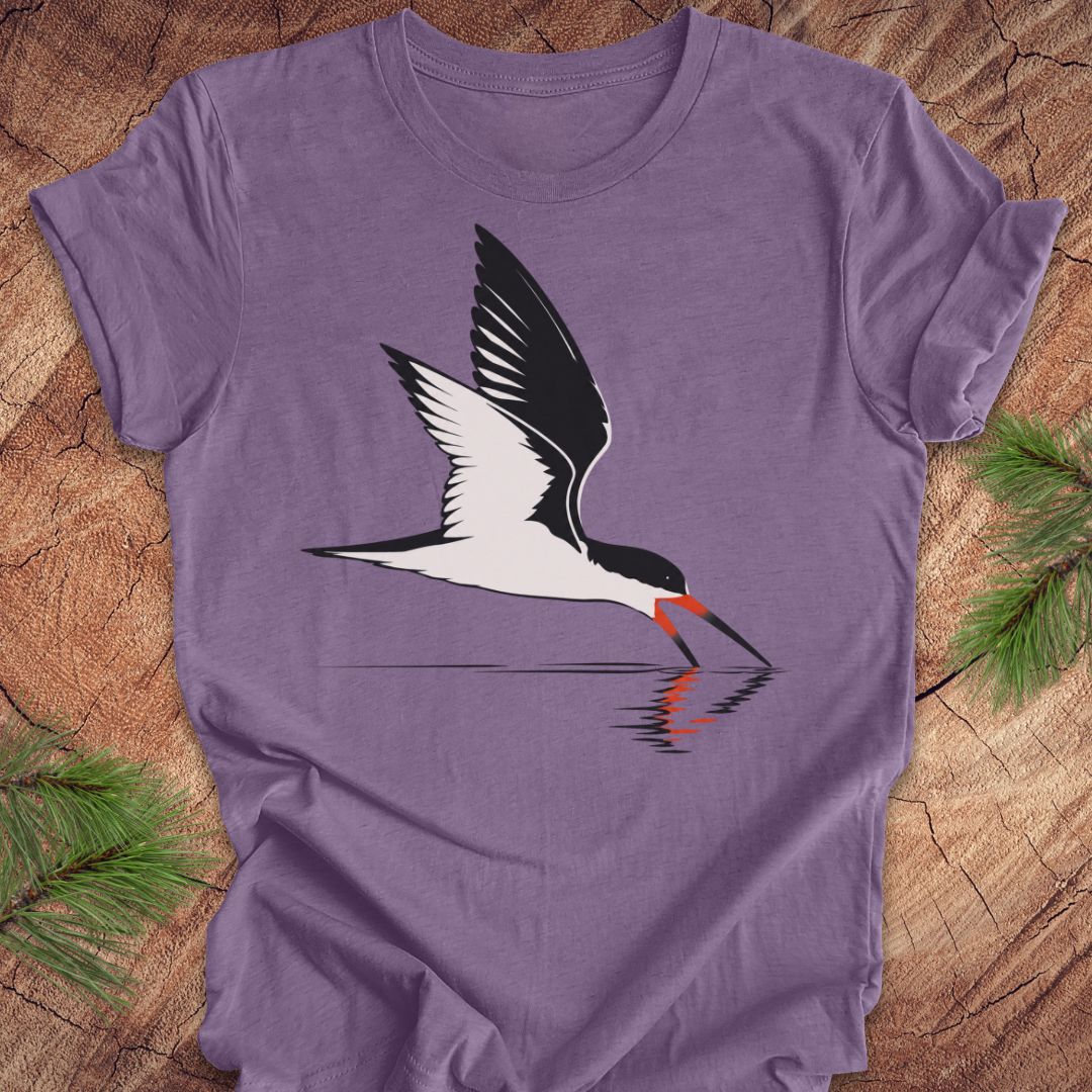 A purple short-sleeve crewneck t-shirt featuring a graphic of a black and white skimmer bird with a red-orange beak, shown flat on a rustic wood background with pine branches.