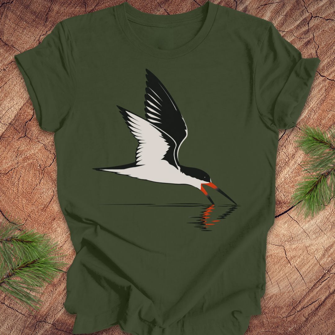 An olive green short-sleeve t-shirt featuring a stylized graphic of a black and white skimmer bird with an orange beak, laid flat on a rustic wood background with pine branches in the corners.