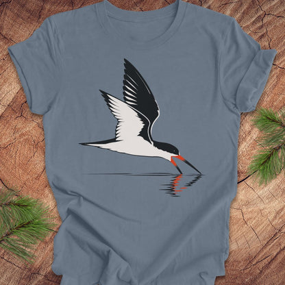 A slate blue crewneck t-shirt featuring a graphic of a black and white skimmer bird with a red-orange beak, lying flat on a rustic wood background with pine branches in the corners.