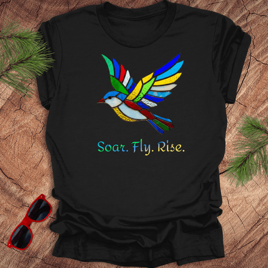 Black t-shirt with colorful bird design and 'Soar. Fly. Rise.' text on a wooden surface with sunglasses and pine branches.
