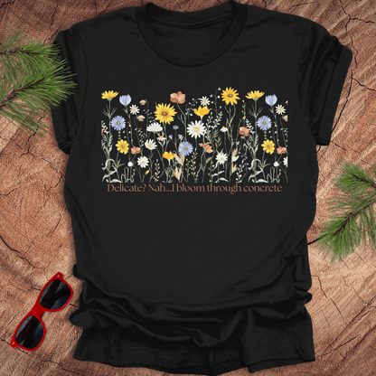 Black t-shirt with floral design and text "delicate? nah. i bloom through concrete.' on a wooden surface.