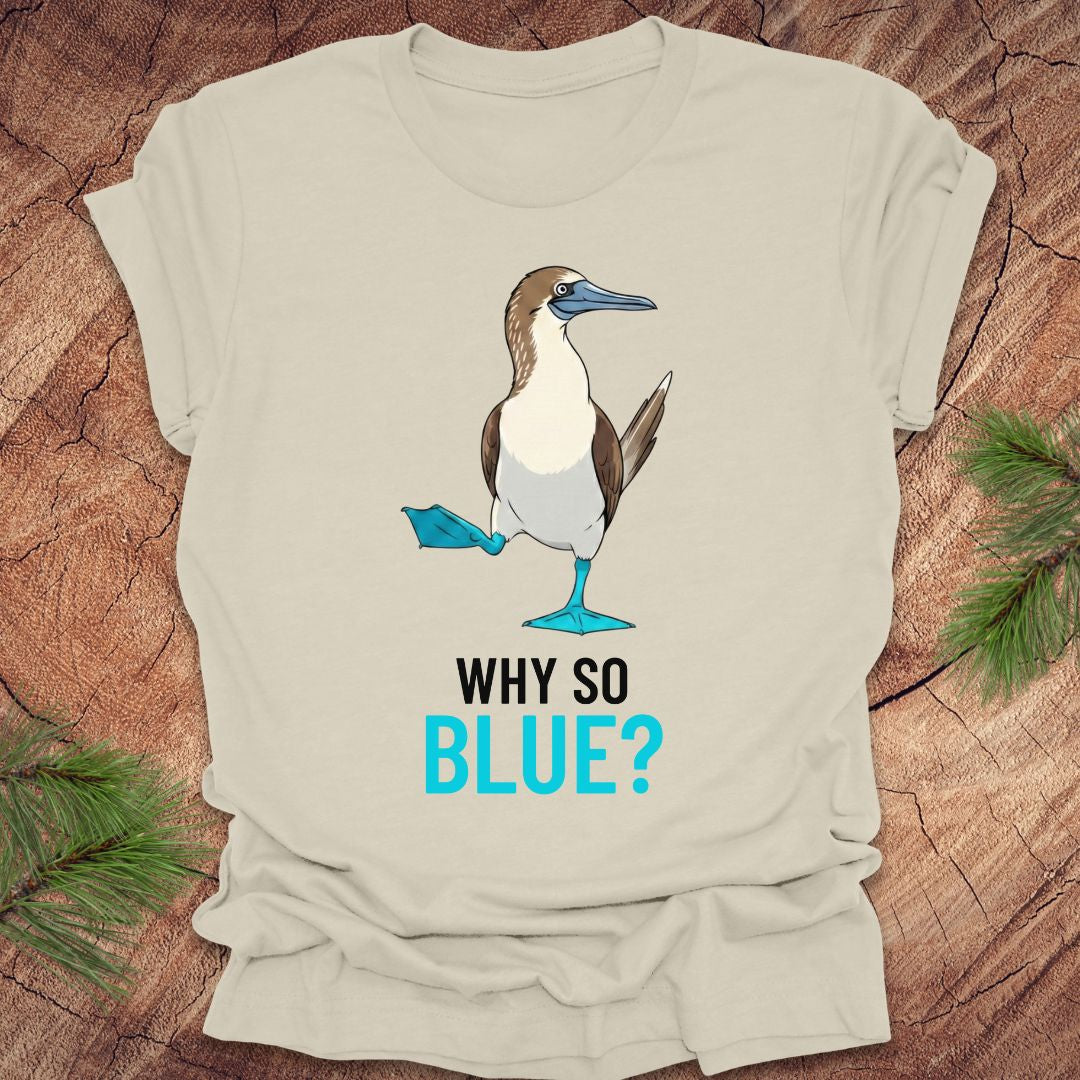 Beige t-shirt with a blue-footed booby bird graphic and 'Why So Blue?' text on a wooden surface.