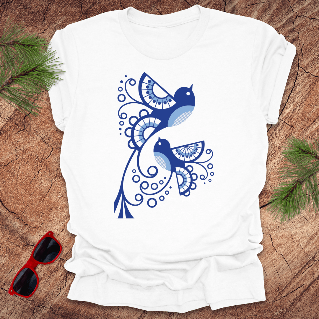 White t-shirt with blue bird design on a wooden surface with sunglasses and greenery.