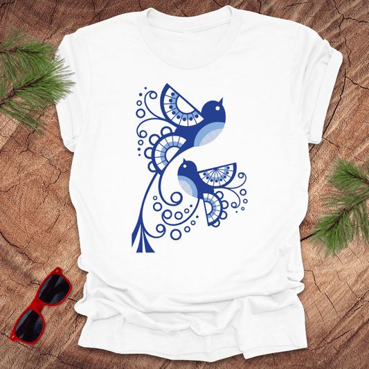 White t-shirt with blue bird design on a wooden surface with sunglasses and greenery.
