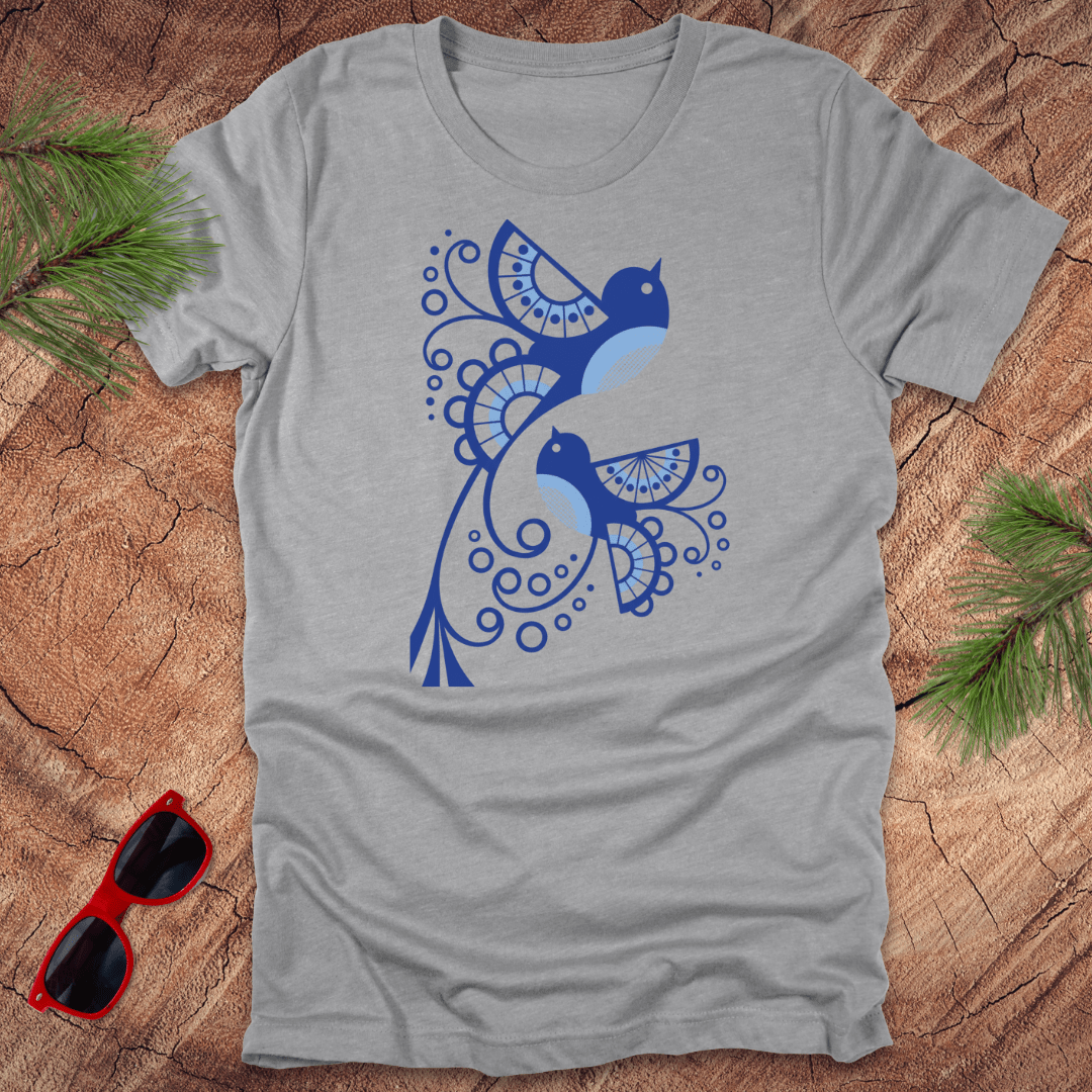 Gray t-shirt with blue abstract bird design on a wooden surface with sunglasses and greenery.