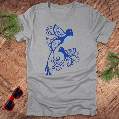 Gray t-shirt with blue abstract bird design on a wooden surface with sunglasses and greenery.