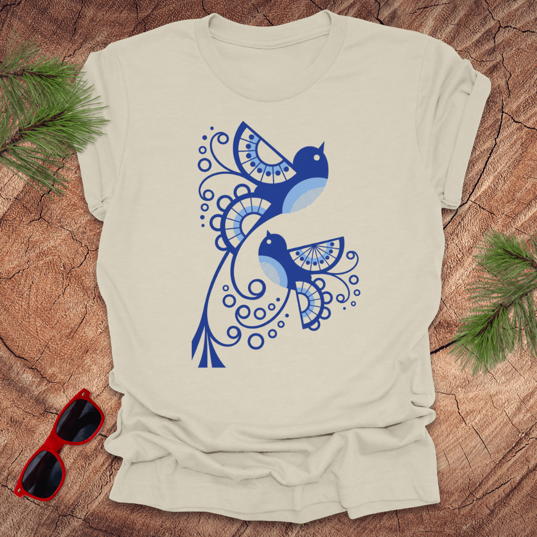 Beige t-shirt with blue bird design on a wooden surface with sunglasses and greenery.