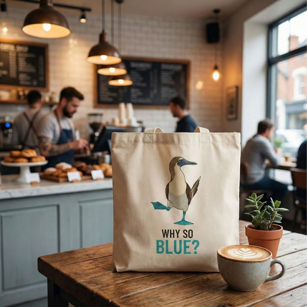 Tote bag with a blue-footed booby bird design and text 'Why So Blue?' on a table in a coffee shop.