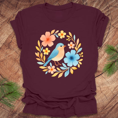 Maroon t-shirt with a colorful bird and floral design on a wooden surface.