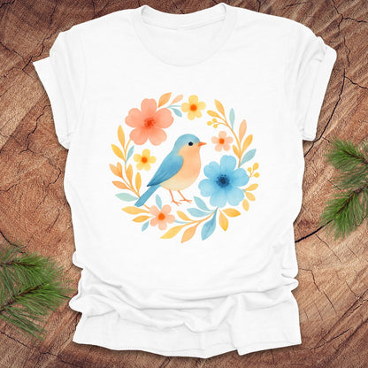 White t-shirt with a colorful bird and floral design on a wooden surface.
