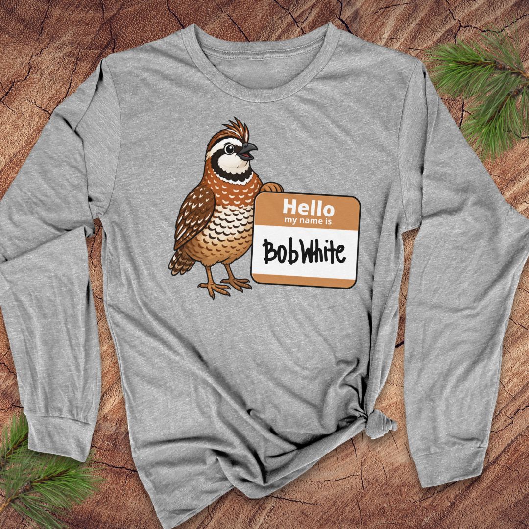 A heather gray long-sleeve t-shirt with a cartoon quail graphic and a 'Hello My Name Is Bob White' nametag, laid flat on a rustic wood background with pine needles.