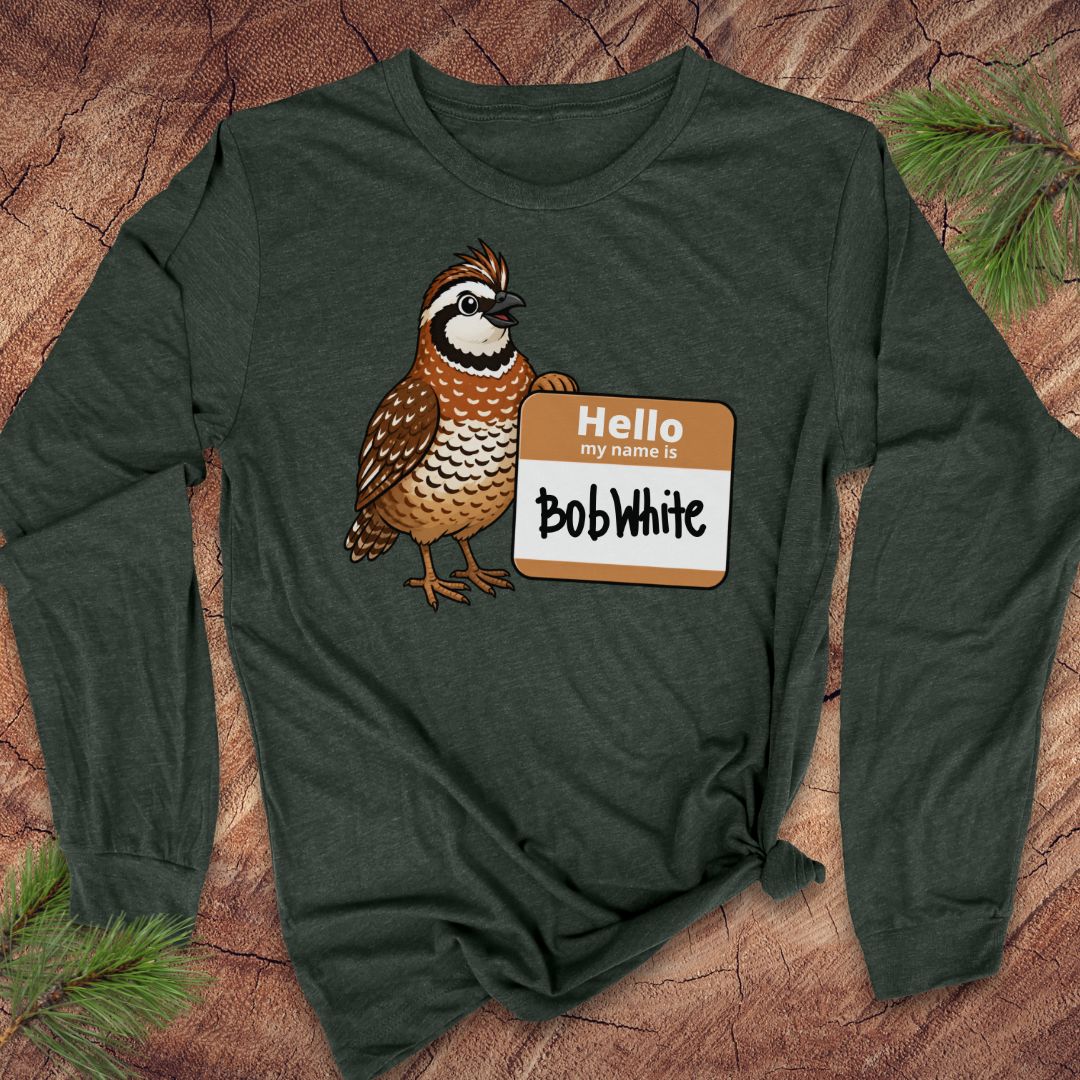 A dark forest green long-sleeve t-shirt with a graphic of a brown and white quail next to a 'Hello My Name Is Bob White' nametag, laid flat on a rustic wood background with pine needles.