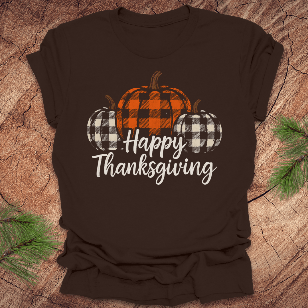 Brown t-shirt with 'Happy Thanksgiving' and checkered pumpkins design on a wooden surface.