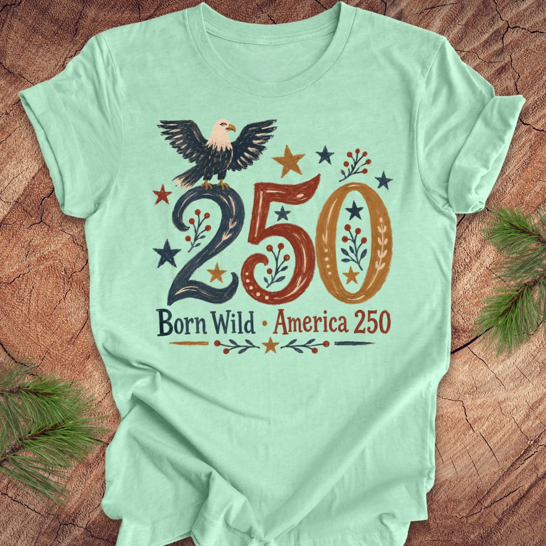 Light green t-shirt with eagle design and 'Born Wild America 250' text on a wooden surface.