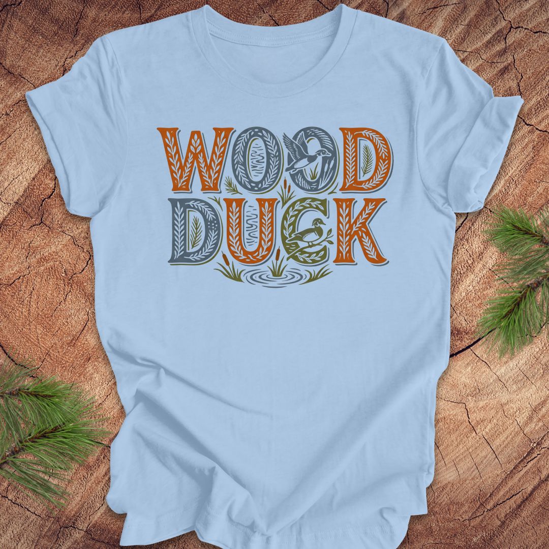 Light blue t-shirt with 'Wood Duck' text design on a wooden surface with greenery.