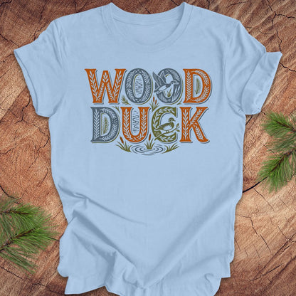 Light blue t-shirt with 'Wood Duck' text design on a wooden surface with greenery.