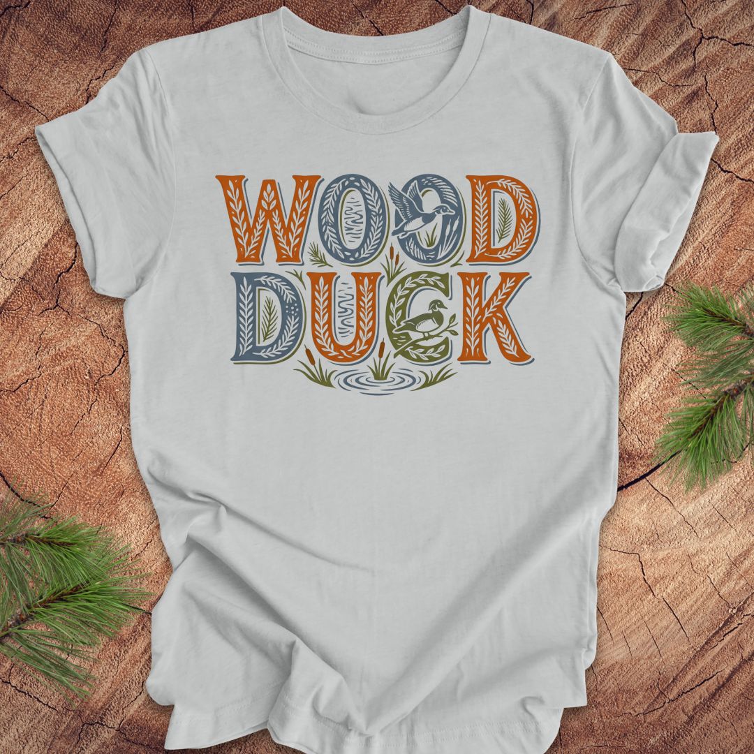 Gray t-shirt with 'WOOD DUCK' text on a wooden surface with greenery.