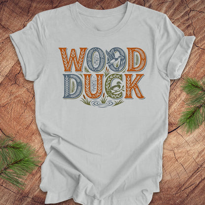 Gray t-shirt with 'WOOD DUCK' text on a wooden surface with greenery.