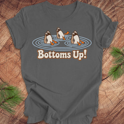 Asphalt gray t-shirt with three Mallards dabbling bottoms-up in rippling pond water above the words Bottoms Up!, laid flat on a rustic wood background with pine branches.