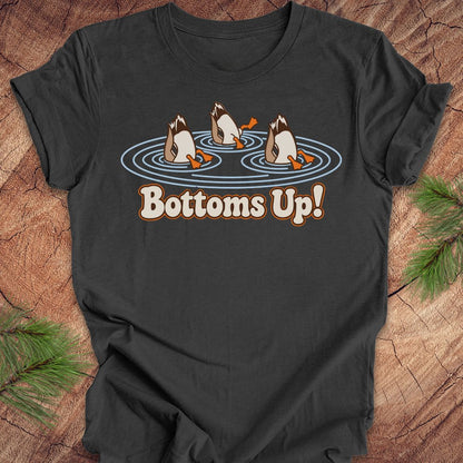 Dark grey heather t-shirt with three Mallards dabbling bottoms-up in rippling pond water above the words Bottoms Up!, laid flat on a rustic wood background with pine branches.