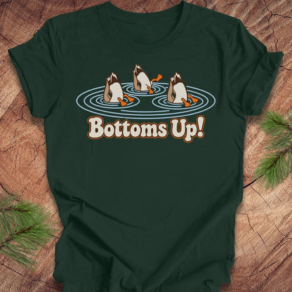 Forest green t-shirt with three Mallards dabbling bottoms-up in rippling pond water above the words Bottoms Up!, laid flat on a rustic wood background with pine branches.