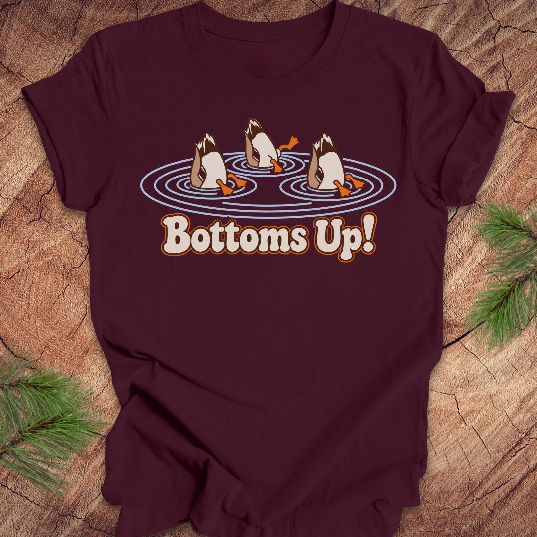 Maroon t-shirt with three Mallards dabbling bottoms-up in rippling pond water above the words Bottoms Up!, laid flat on a rustic wood background with pine branches.