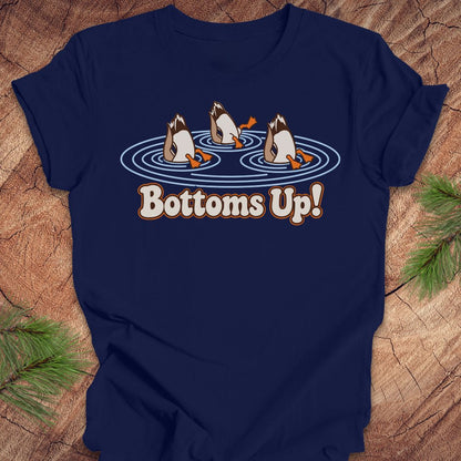 Navy t-shirt with three Mallards dabbling bottoms-up in rippling pond water above the words Bottoms Up!, laid flat on a rustic wood background with pine branches.