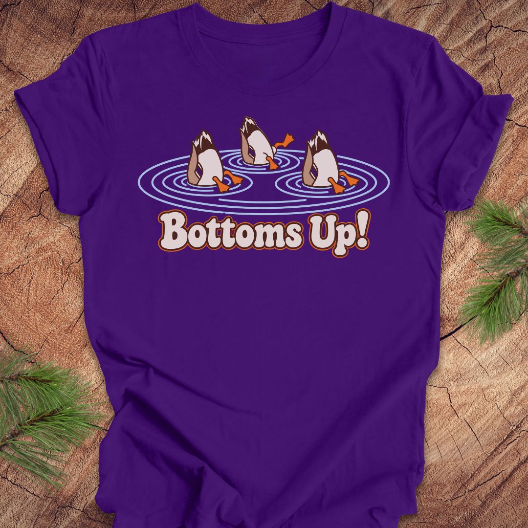 Purple t-shirt with three Mallards dabbling bottoms-up in rippling pond water above the words Bottoms Up!, laid flat on a rustic wood background with pine branches.