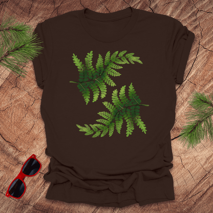Brown t-shirt with green leaf design on a wooden surface with sunglasses and pine branches.