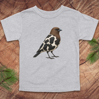 A light heather gray short-sleeve t-shirt featuring a detailed graphic of a brown and black-and-white speckled bird on the chest, laid flat on a rustic wood slice background with pine needles in the corners.
