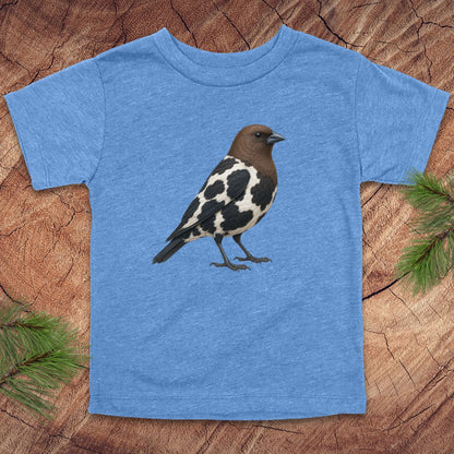 A heather blue short-sleeve t-shirt featuring a graphic print of a brown and black-and-white speckled bird, laid flat on a rustic wood slice background with pine needles in the corners.