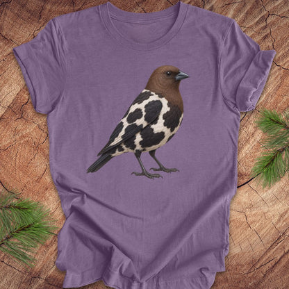 A purple short-sleeve t-shirt featuring a detailed graphic of a brown and black-and-white speckled bird, laid flat on a rustic wood slice background with pine needles in the corners.