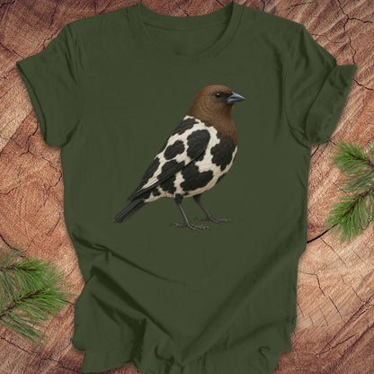 An olive green short-sleeve t-shirt featuring a detailed graphic of a bird with a brown head, black and white spotted body, and dark legs, laid flat on a rustic wood background with pine branches.