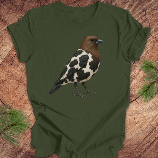 An olive green short-sleeve t-shirt featuring a detailed graphic of a bird with a brown head, black and white spotted body, and dark legs, laid flat on a rustic wood background with pine branches.