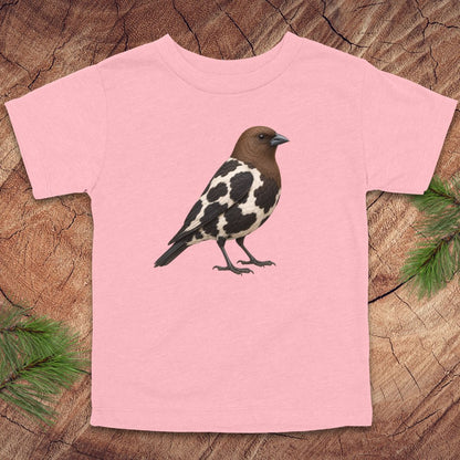 A light pink short-sleeve t-shirt featuring a graphic of a mottled brown, black, and white bird, displayed flat on a rustic wood slice background with pine needles in the corners.